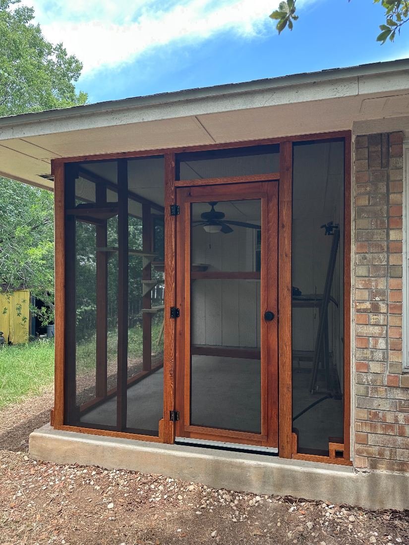 Hand-built cedar screen door with a real doorknob and black hinges, set into a finished screened porch catio with climbing shelves inside